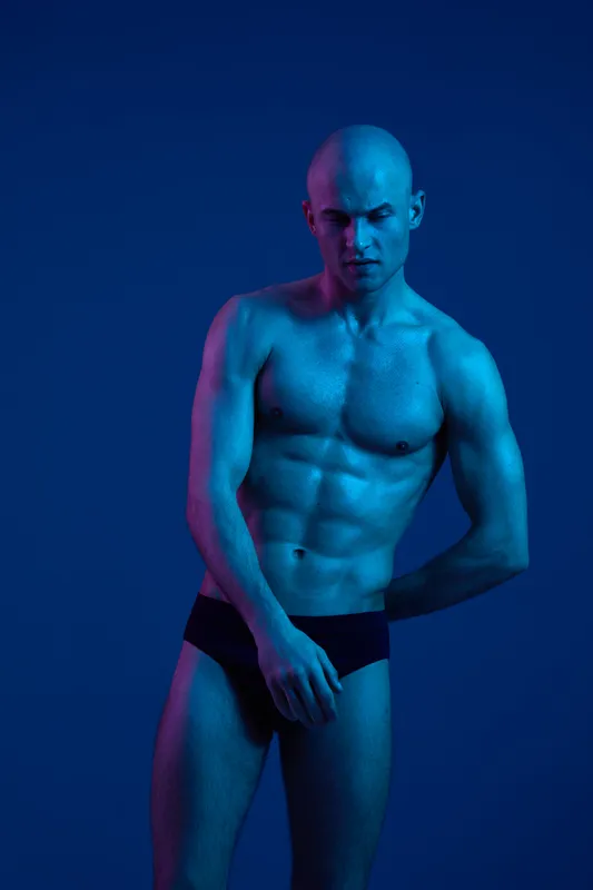 Athletic man posing in blue lighting, showcasing strong physique and confident expression. The vibrant colors emphasize strength and artistic composition.