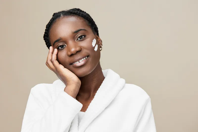 Young African woman applying skincare cream, smiling, dressed in a white robe, with natural makeup against a neutral background, promoting self care and beauty