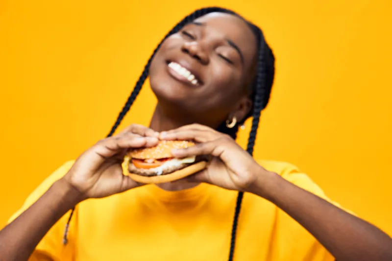 Joyful young Black woman enjoying a delicious hamburger in a vibrant yellow setting, expressing happiness and satisfaction with her meal