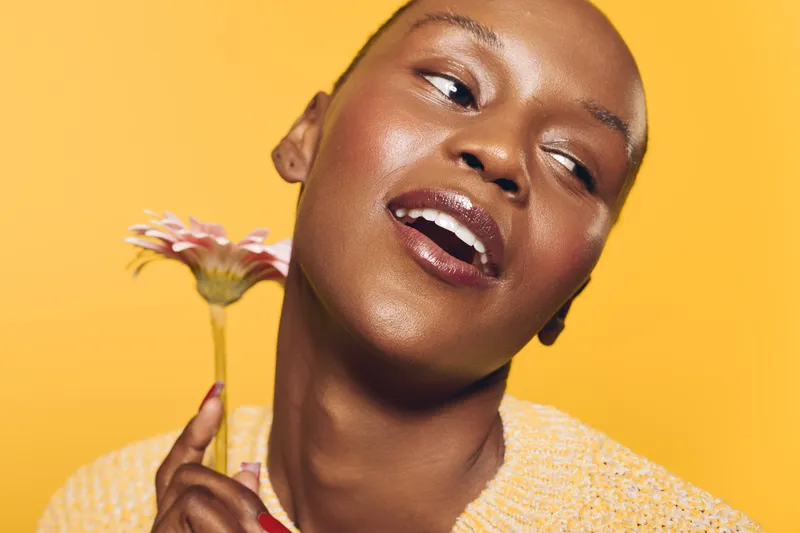 Close-up of happy dark skinned African woman holding flower near face, smiling with eyes looking aside on yellow background. Concept of beauty, joy, and natural skincare.