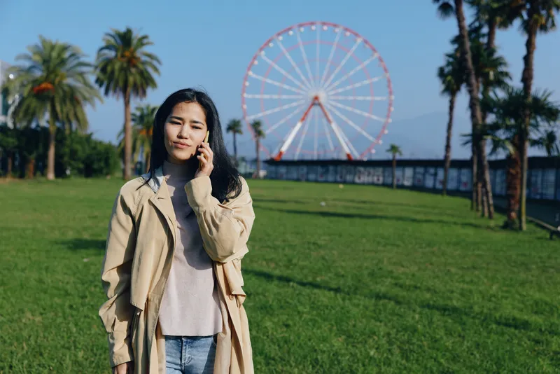 Woman in beige coat standing in a green park with a distant ferris wheel, casual wardrobe, sunny day, relaxed pose, travel vibe, lifestyle photography