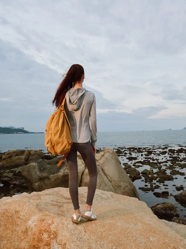 Young woman with backpack enjoying serenity by the sea during a cloudy day, wearing casual athletic clothing, representing adventure and freedom