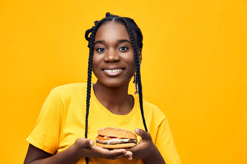 Young Black woman smiling while holding a delicious burger against a vibrant yellow background, showcasing happiness and joy in healthy eating