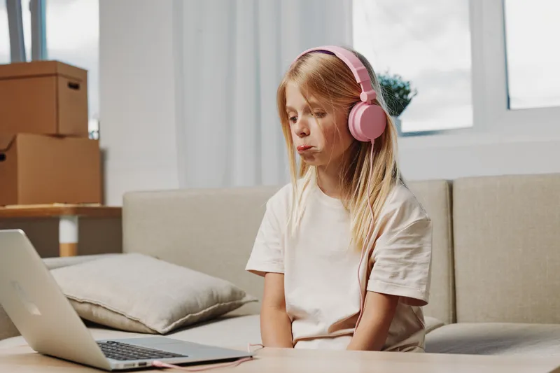 A young girl with long hair wearing pink headphones focuses on a laptop screen in a cozy living room with natural light and cardboard boxes.