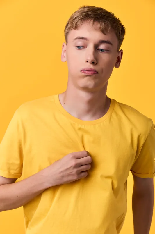 Thinking young man in a yellow shirt stands in a studio, reflective mood, casual fashion, bright lighting, monochrome yellow backdrop, subtle pose and calm expression.