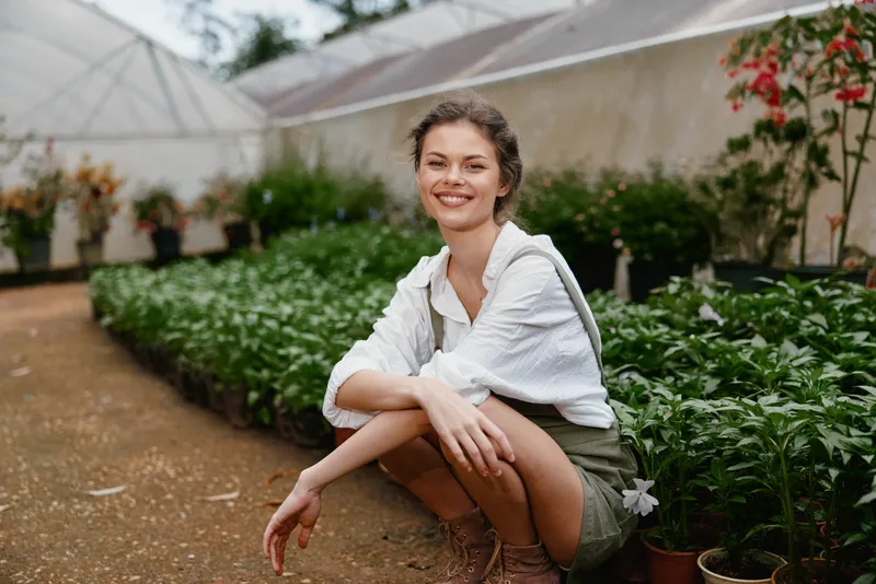 Young woman smiling in a greenhouse, wearing a white shirt and green overalls, surrounded by vibrant plants, conveying a refreshing and natural atmosphere