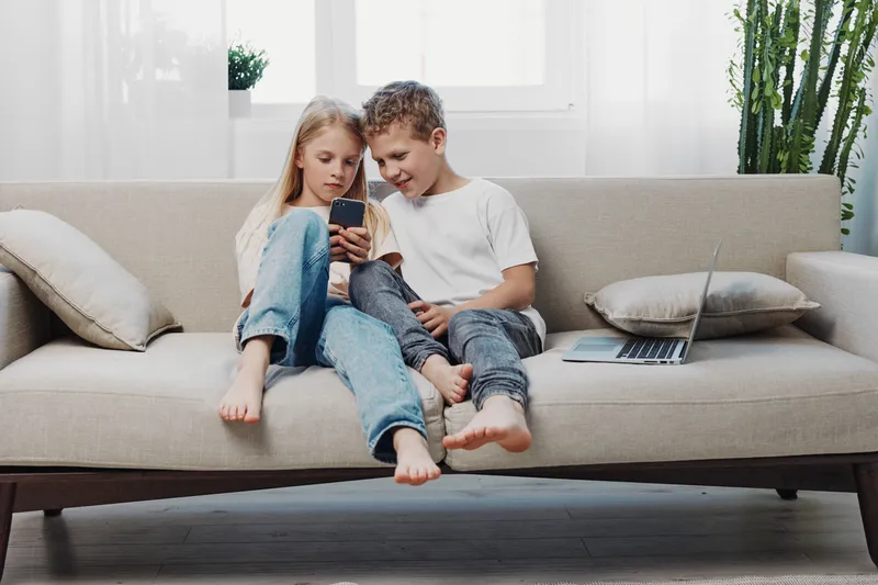 Two children sit barefoot on a sofa looking at a smartphone together in a bright room with a laptop nearby. Concept of technology, childhood, and casual home life.