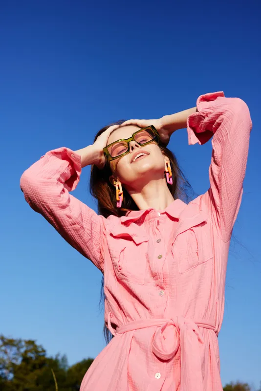 Portrait of a fashionable young woman outdoors in a pink dress against a clear blue sky, expressing joy with stylish sunglasses, ideal for showcasing summer fashion trends