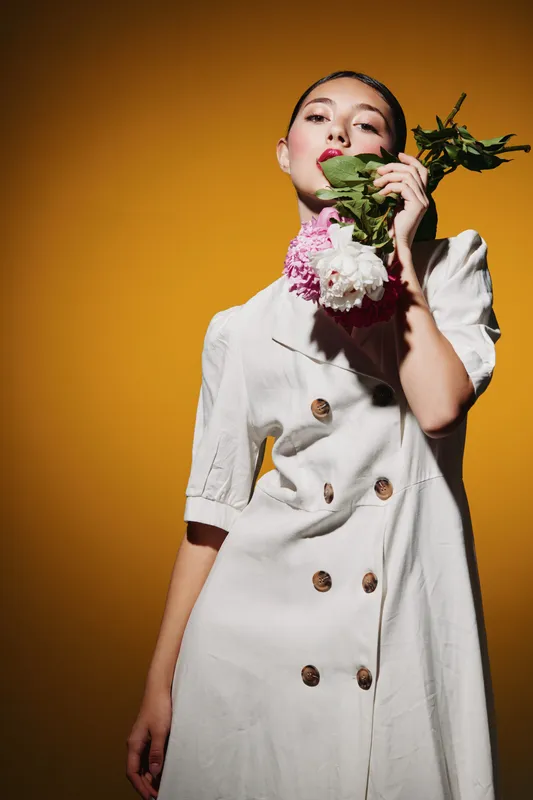 Young woman in a white buttoned dress poses holding pink flowers on a yellow background. Creative beauty fashion portrait with vivid colors and confident expression in studio light.