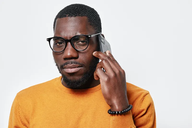 African American man with dark skin wearing glasses and orange sweater talking on smartphone with serious expression isolated on white background in studio portrait.