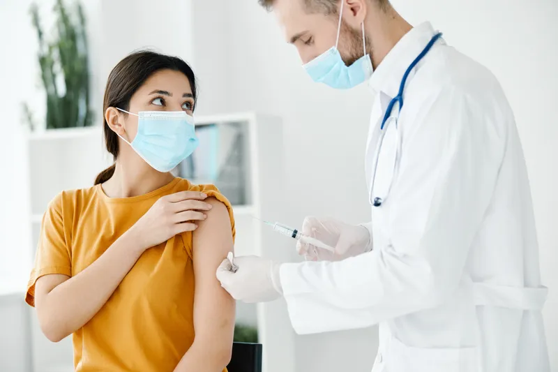 Пациент в медицинской маске смотрит на врача со шприцем в руке, вакцинация