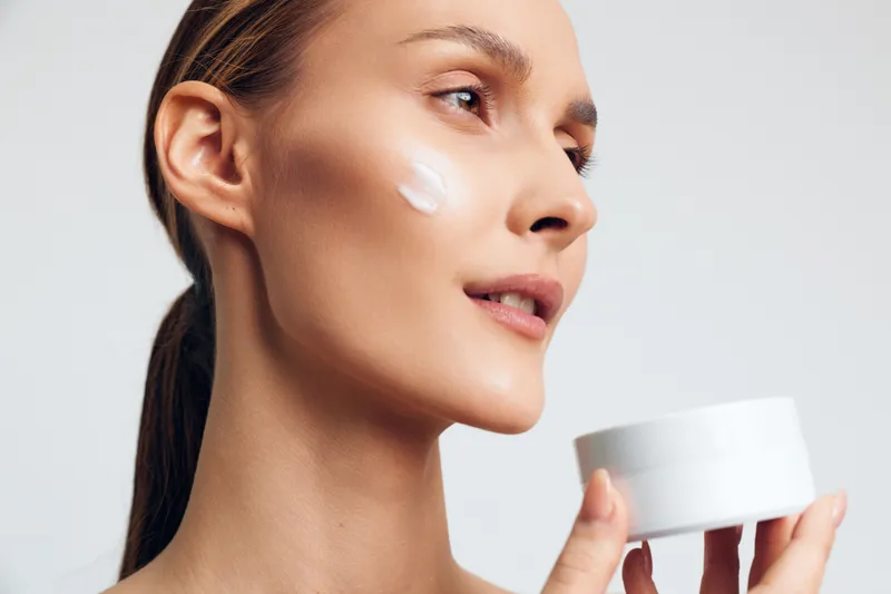 Young woman with clear skin applying white face cream on cheek holding container close-up on white background. Concept of skincare, beauty routine, wellness, and self-care.
