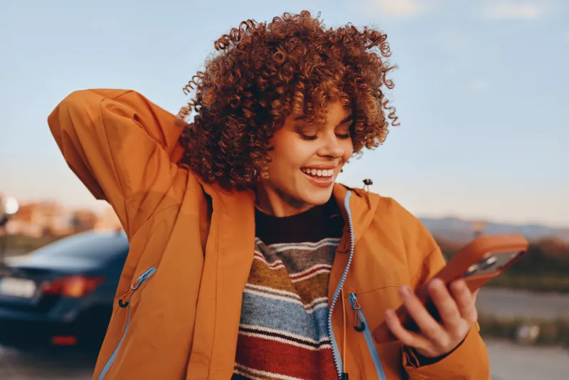 Smiling woman lifestyle outdoors, curly hair under orange jacket, holding phone and scrolling, warm sunset light and city road with cars in background, casual communication moment