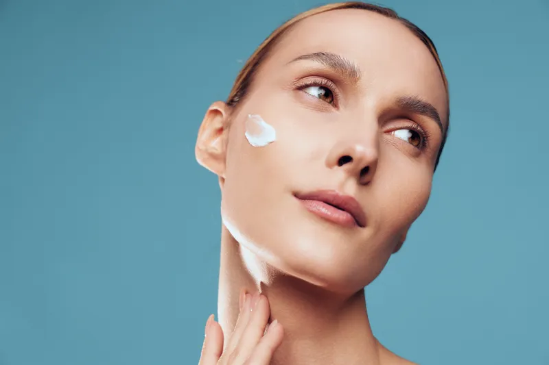 Young woman applying white face cream on her cheek with fingers. Close-up portrait on blue background. Concept of skincare, beauty routine, and healthy skin care.