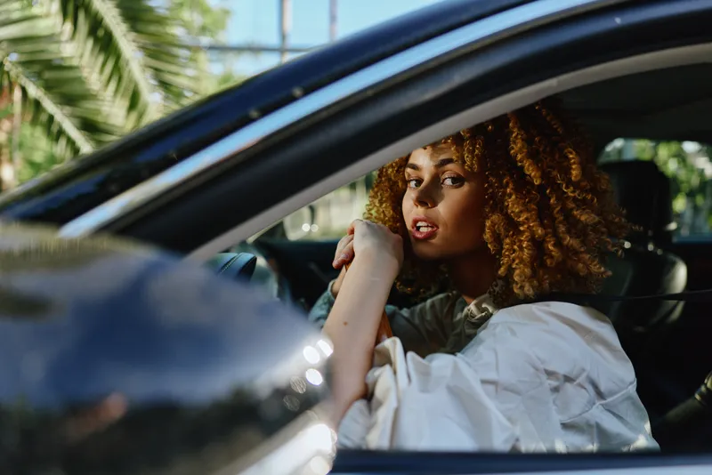 Afro woman with curly hair driving car outdoors, wearing white shirt, natural light, relaxed expression, green palm trees background, casual style.
