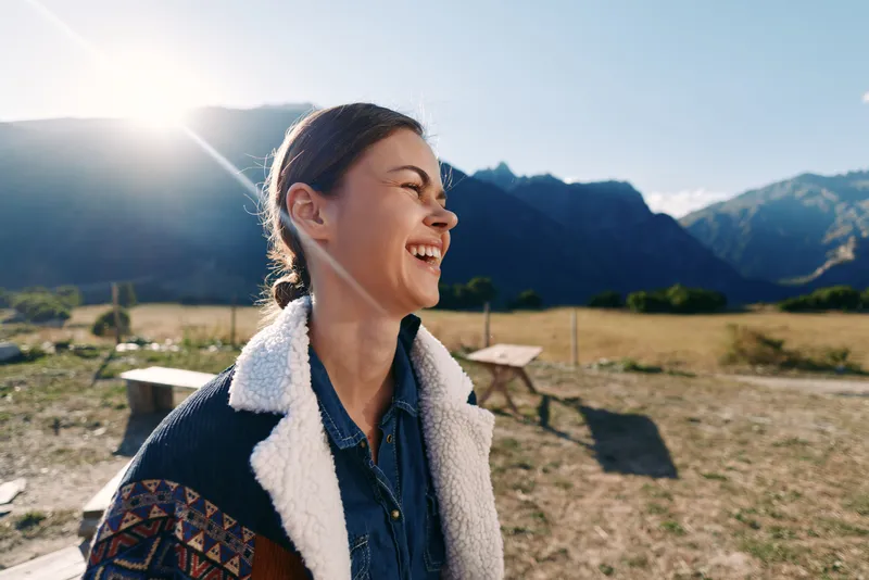 Woman smile laughing outdoors in sunlight with mountains backdrop, wearing warm jacket and denim shirt, candid portrait enjoying nature in a meadow by picnic tables and open field.
