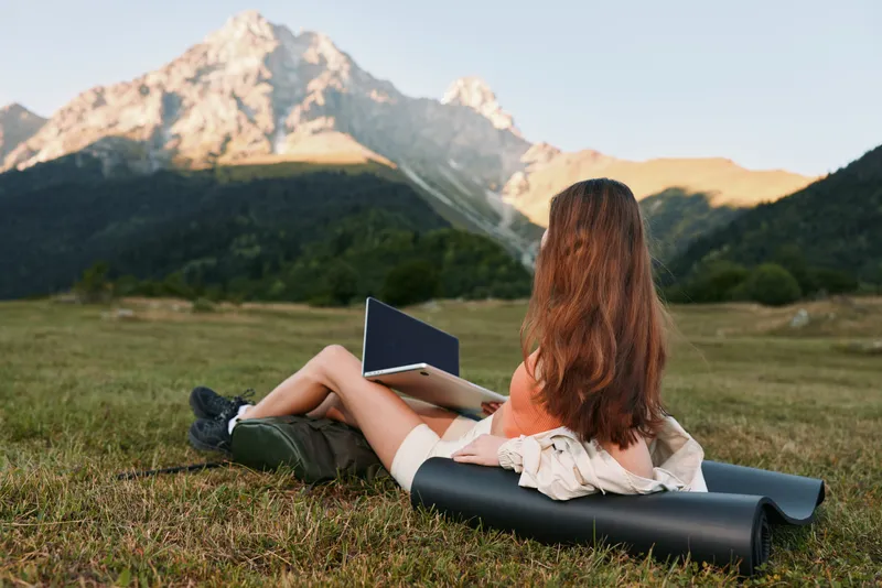 A woman sits on a yoga mat outdoors with a laptop, in a grassy meadow under mountain light. She wears casual clothes, appears focused yet relaxed, blending nature and productivity.