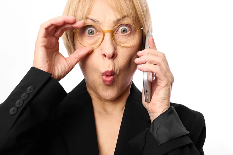 Woman glasses phone surprised older business portrait studio adjusting frames, holding phone to ear and widening eyes, professional suit, white background, bright light