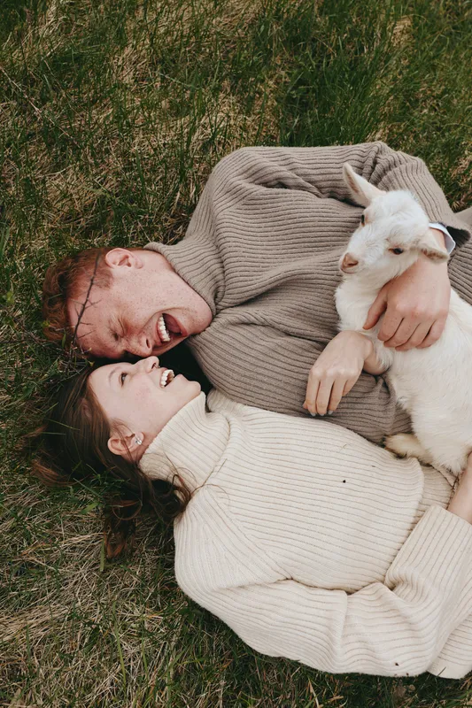 A couple lying on grass in cozy sweaters, smiling, while a white goat kid rests beside them, capturing a warm lifestyle moment of affection, nature, and playful bonding outdoors.