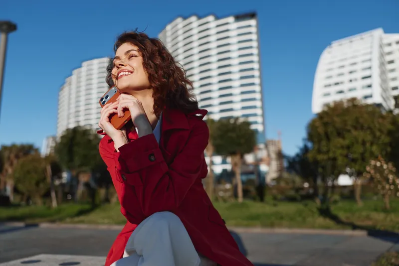 Smiling woman with curly hair talking on a smartphone outdoors, wearing a red coat. Urban park with modern buildings in the background. Energetic, confident lifestyle moment.
