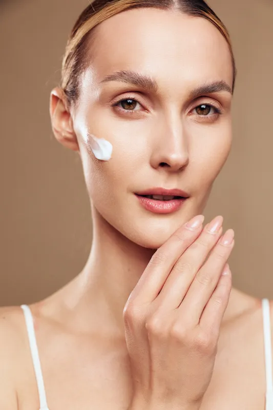 A young woman with clear skin applies white cream on her cheek, looking directly at the camera against a beige background. Concept for skincare, beauty, and natural care.