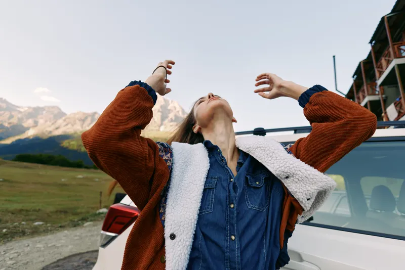 Woman in warm shearling jacket stands by car with arms raised, enjoying mountains during travel roadtrip, expressive moment of freedom and joy on a scenic countryside drive.