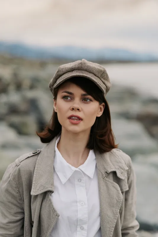 Young woman wearing a vintage cap and light jacket stands outdoors near a rocky shore with blurred water and mountains in the background. Concept of autumn fashion and nature.