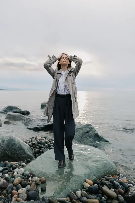 Young woman in casual clothing stands on rocky shore with hands on head, eyes closed, facing calm sea under cloudy sky. Concept of relaxation, contemplation, and nature connection.