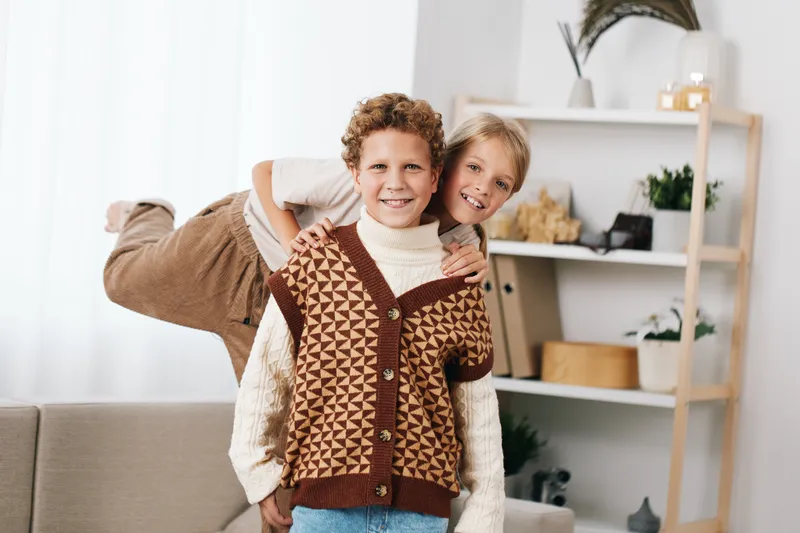 Two smiling children hug playfully indoors, wearing knitted sweaters and warm tones, capturing a joyful family moment and candid friendship in a bright living room.