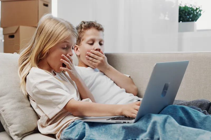 Two children sitting on a sofa, yawning while using a laptop. Cozy home environment with natural light from window, concept of tiredness, screen time, and childhood relaxation.