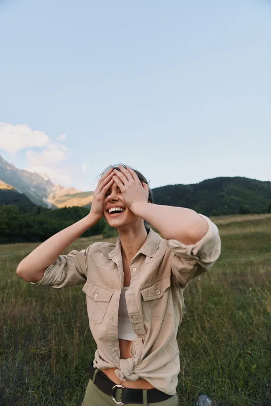 Young woman in a beige shirt laughs and covers her eyes in a grassy field with mountains in the background