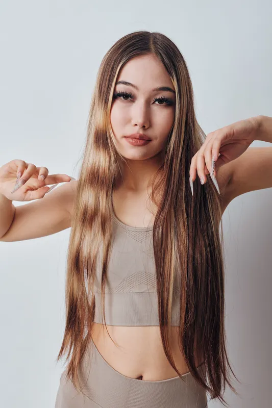 Young woman with long brown hair posing in beige crop top and pants showing confident expression and hand gestures against a plain background in studio light.