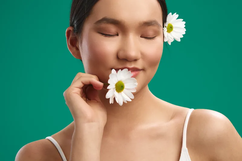 asian woman with a gentle smile holds a daisy near her lips in a simple studio portrait, creating a serene, natural vibe and a warm, approachable mood.