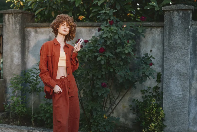 A stylish woman poses outdoors in an orange coordinated outfit, leaning against a garden wall with red blooms. Casual fashion portrait for everyday lifestyle and confidence