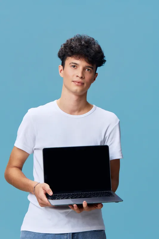 young man holding laptop with blank screen standing against blue background wearing casual white t-shirt