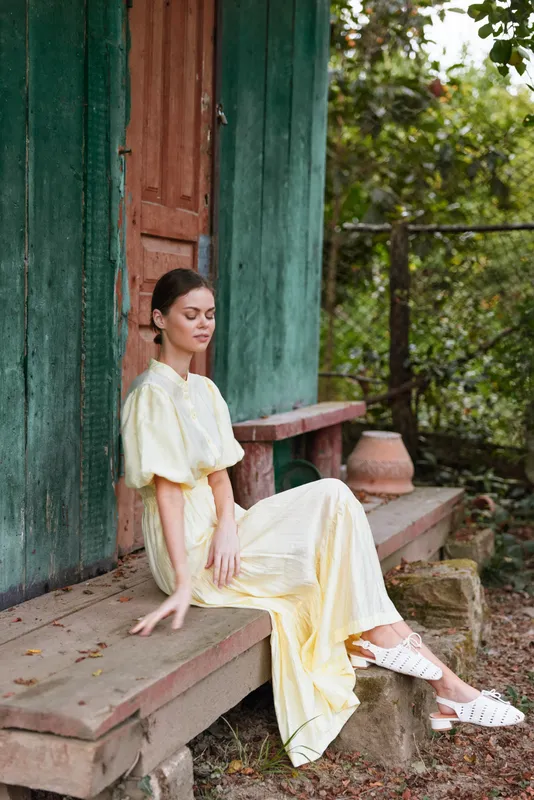Woman in a pale yellow dress sits on a wooden porch beside a green door, relaxed and contemplative in a rustic outdoor setting, sunlight creates a serene countryside mood.