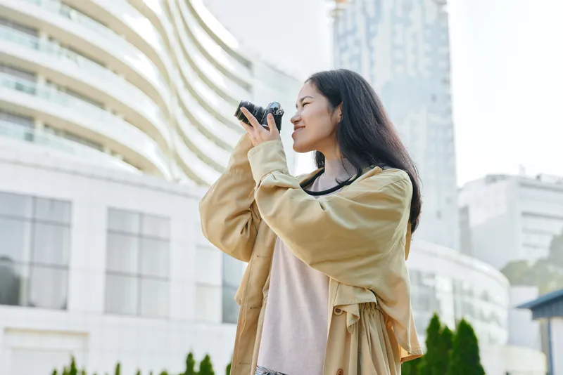 Woman in a beige coat photographs the city skyline with a smartphone in an urban plaza, smiling as modern buildings rise behind her during daylight and urban energy
