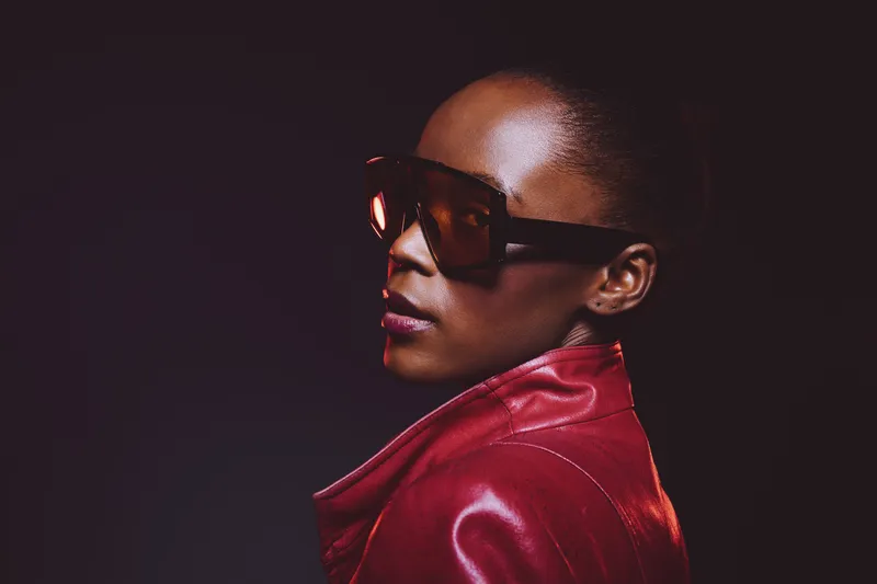 Stylish African American woman with dark skin wearing large sunglasses and a red leather jacket poses confidently against a dark background. Concept of fashion and elegance.