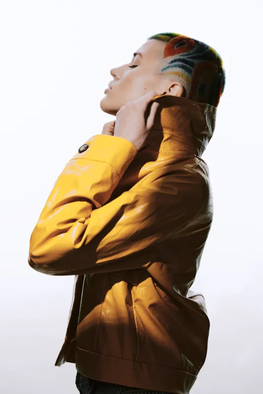 Young woman with colorful shaved hair adjusts her vibrant yellow jacket against a bright white background, showcasing individuality and bold fashion choices.