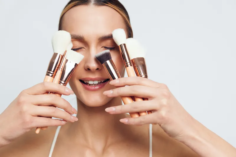 Smiling woman holding multiple makeup brushes close to her face with eyes closed on white background. Concept of beauty tools, cosmetics, and skincare routine with natural lighting.