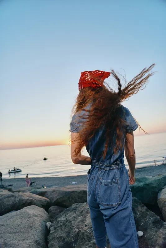 lifestyle woman in modern boho-western denim street style with red bandana stands by the rocky beach at sunset in natural film color tones