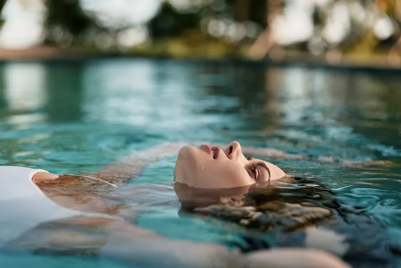 Relaxed swimmer floats in a sunlit pool, head above water, eyes closed, enjoying a peaceful wellness moment, calm atmosphere, casual lifestyle by crystal blue water and gentle light reflecting