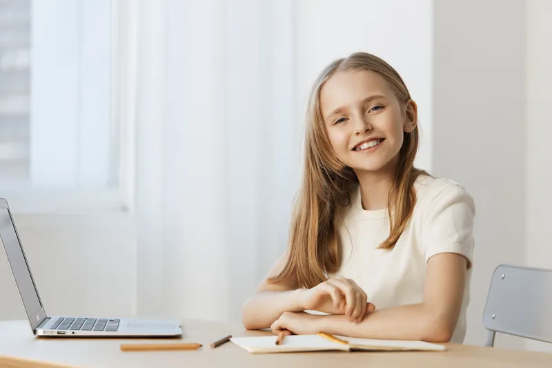 Happy child studying at home, smiling and looking at the camera, wearing a white shirt, sitting at a desk, bright indoor environment with natural light