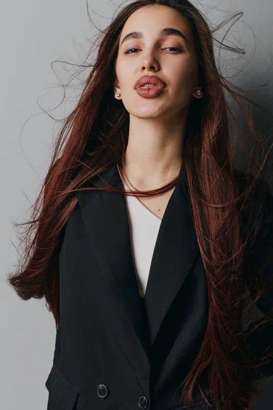 Young woman with long brown hair wearing black blazer and white shirt posing with eyes closed on gray background. Fashion portrait with natural look and relaxed expression indoors.