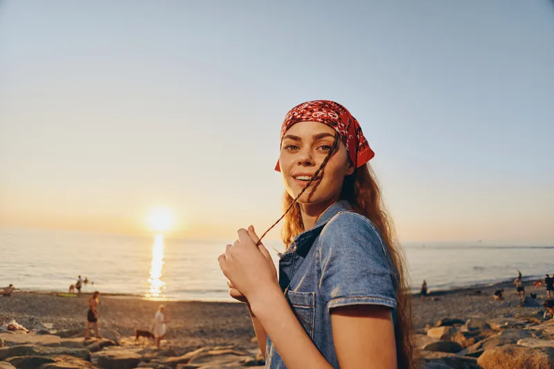 Lifestyle woman in modern boho-western denim street style wearing a red bandana at sunset by the beach, film color with warm natural lighting and relaxed mood.
