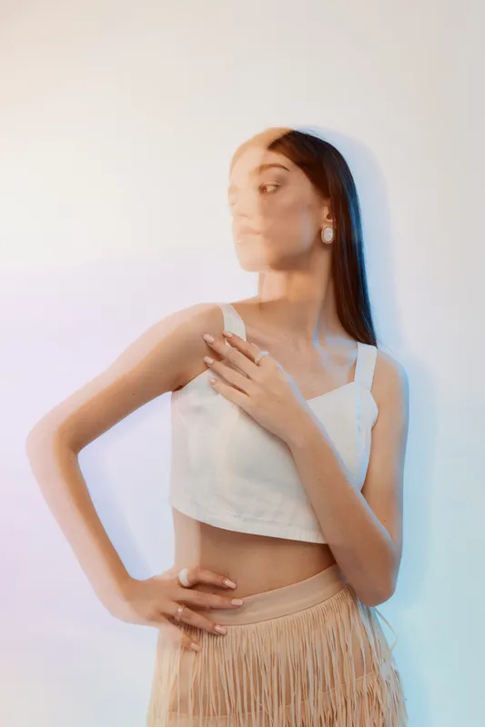 Young woman beauty fashion model poses with hand on chest wearing white top and beige skirt. Soft lighting creates elegant mood on neutral background. Concept of style and confidence.