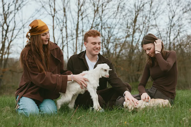 Cheerful group outdoors, smiling around a playful goat kid, sunlit grass, casual outfits, gentle interaction, candid moment, natural colors, rustic setting, warm atmosphere