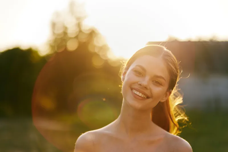 Happy young woman smiling in sunlight, enjoying nature during golden hour, with a warm background and soft colors, representing joy and positivity in life
