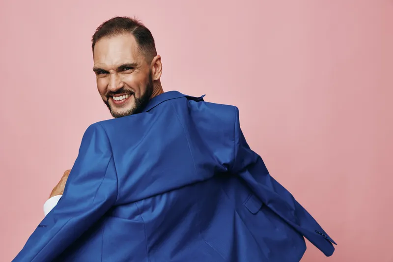 Professional man in a blue suit turns with a confident smile against a solid pink background, isolated colored backdrop highlighting emotion and a dynamic business presence.