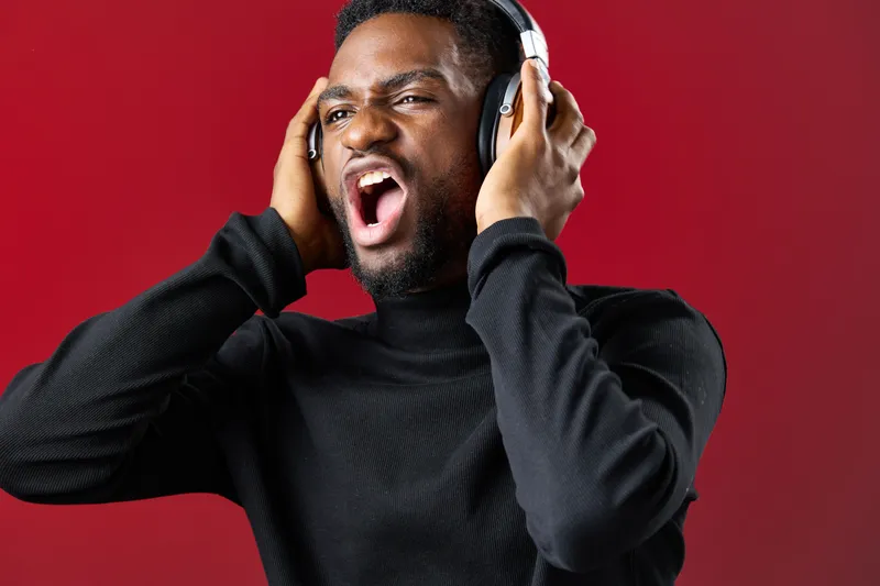 Young African American man passionately singing with headphones against a vibrant red background, expressing joy and excitement through music
